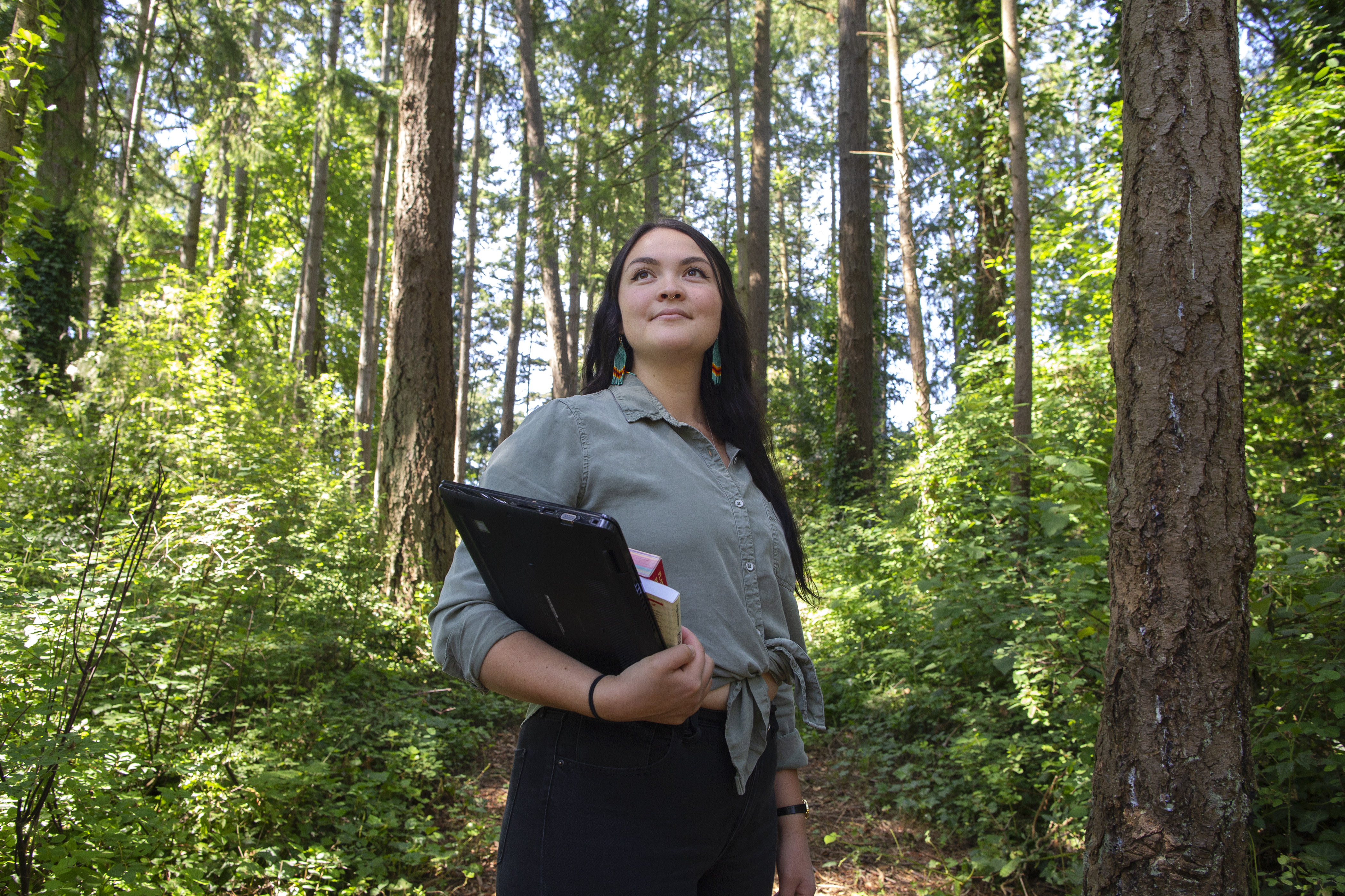A student stands in the middle of a forest surrounded by tall trees and green foliage. The student holds a laptop and books and looks into the distance.