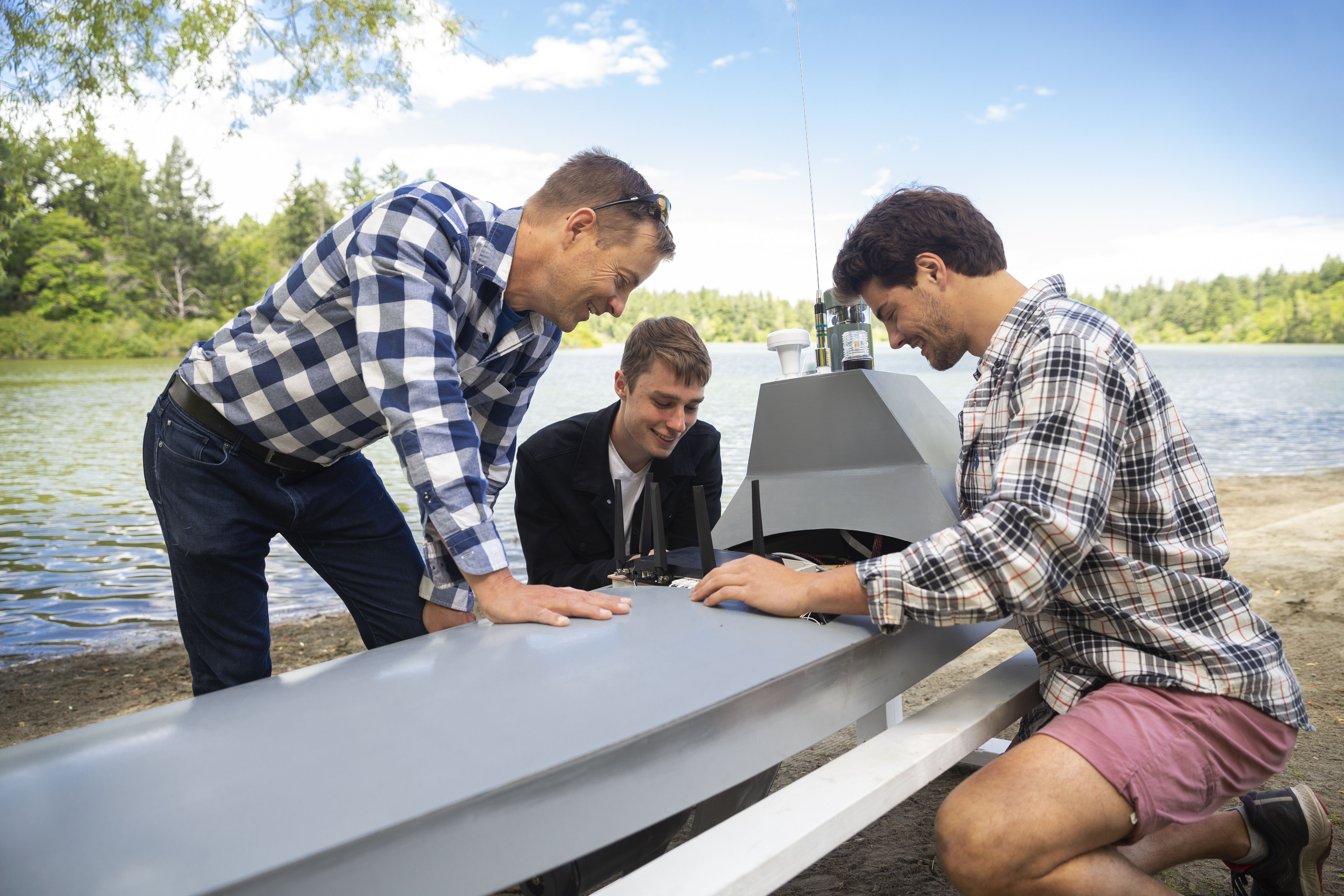 Two students and an employer look at the controls on an autonomous water vehicle that can collect environmental data. The vehicle sits on a small beach and there is water behind it. They are preparing to launch.